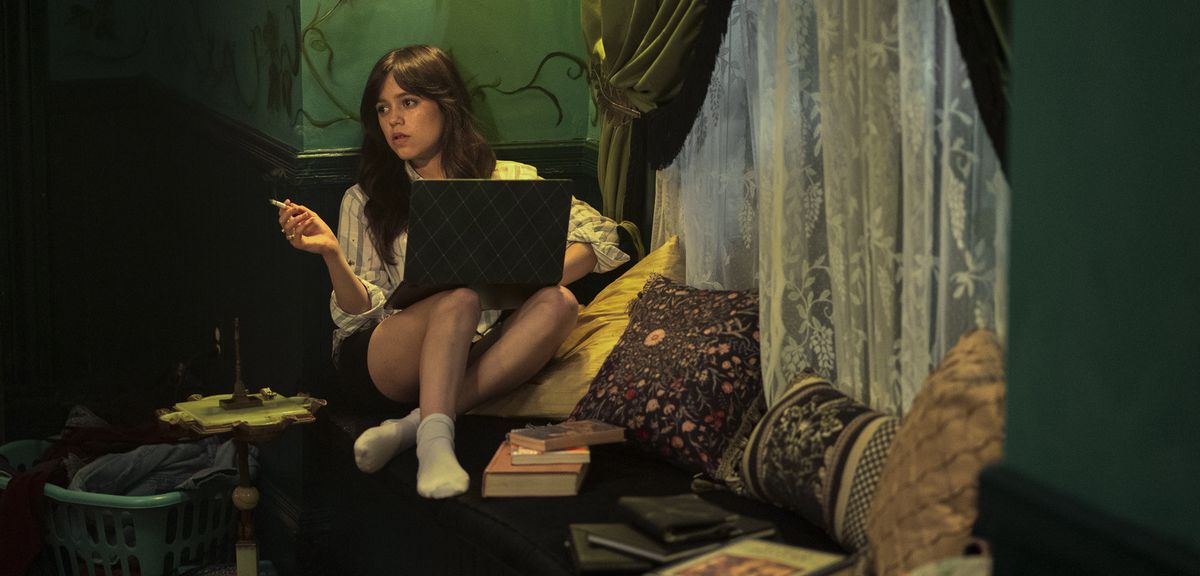 Cairo (Jenna Ortega) sitzt und raucht mit einem Laptop auf dem Schoß und zu einem Stuhl gefalteten Beinen, umgeben von Kissen, Büchern und einem hauchdünnen Spitzenvorhang in „Miller's Girl“.