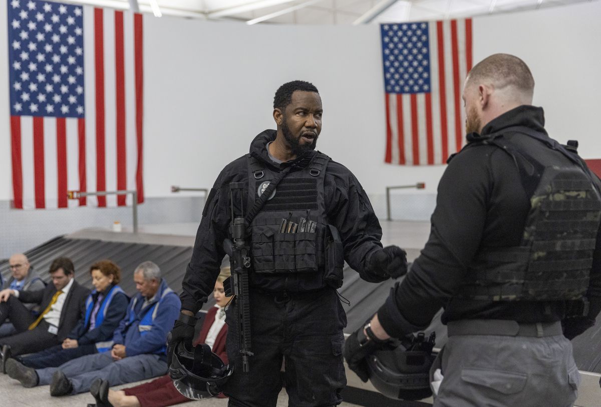 Michael Jai White, con un chaleco antibalas y un rifle colgado del hombro, habla con otro hombre que lleva un chaleco antibalas mientras los rehenes se alinean frente a la cinta de equipaje del aeropuerto en One More Shot.