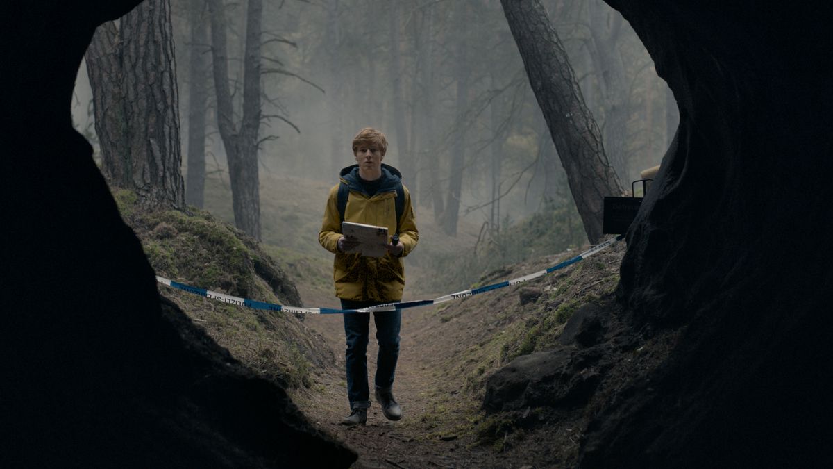 Un joven vestido con un impermeable amarillo se encuentra frente a la entrada dividida de una gran cueva en un bosque en la oscuridad.