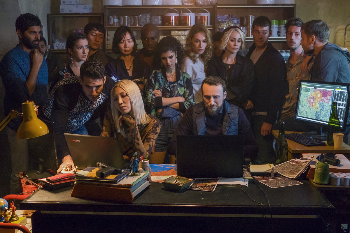 Een groep mensen, waaronder de hoofdrolspelers van Sense8, die over de schouder van twee mensen staat te typen op laptops in Sense8.
