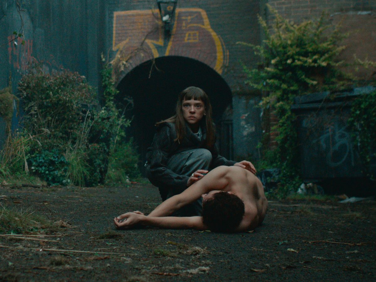 Shira Haas als DS Maplewood knielt naast een lichaam op een binnenplaats vol met planten in Bodies.