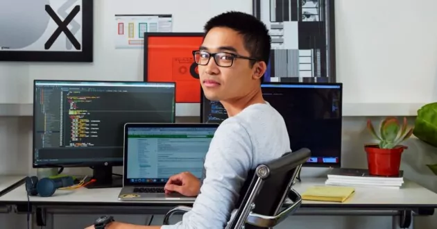 Jonge persoon met een bril zit in een bureaustoel voor drie computerschermen, kijkt naar de camera en glimlacht