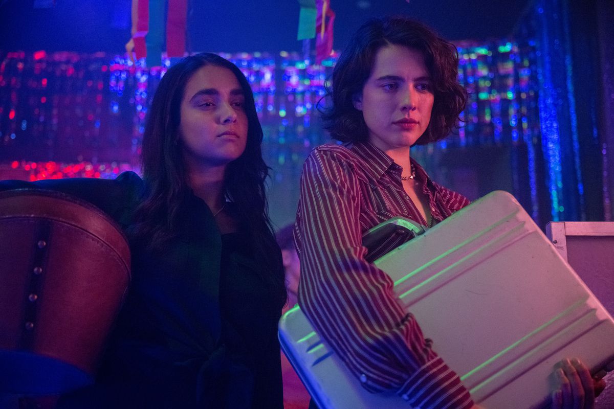 Jamie ve Marian, Drive-Away Dolls filminden bir sahnede, biseksüel ışıklarla dolu bir barda bir evrak çantasını yakına tutuyorlar.
