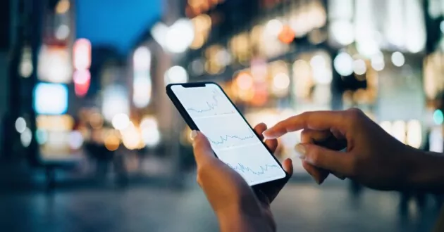 ライトアップされた都市の高層ビルを背景にダウンタウンの街路でスマートフォンで金融取引データを読む人