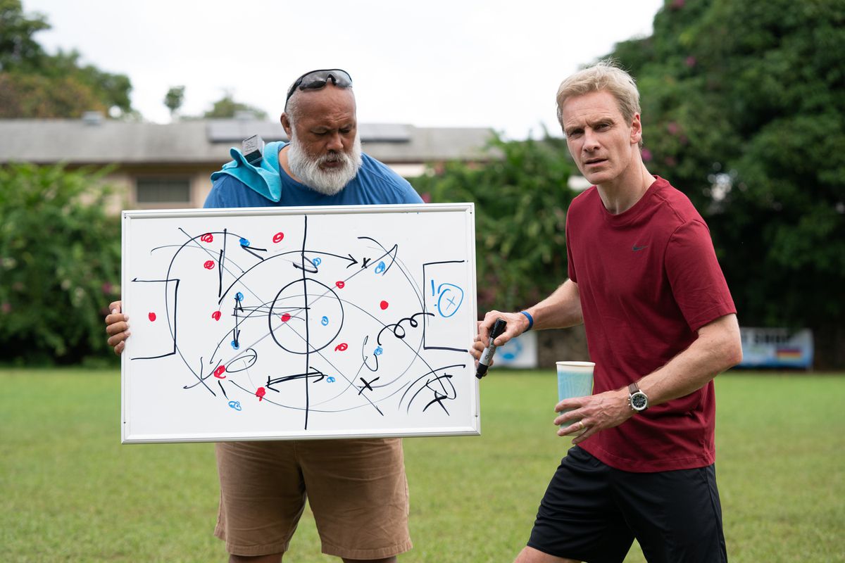 Ace (David Fane) hält ein Whiteboard in der Hand, während Trainer Thomas Rongen (Michael Fassbender) in „Next Goal Wins“ abseits des Bildschirms sein Team belehrt.