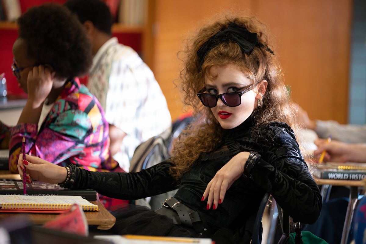 Kathryn Newton, büyük saçlı, Lisa Frankenstein'da okul masasında oturuyor