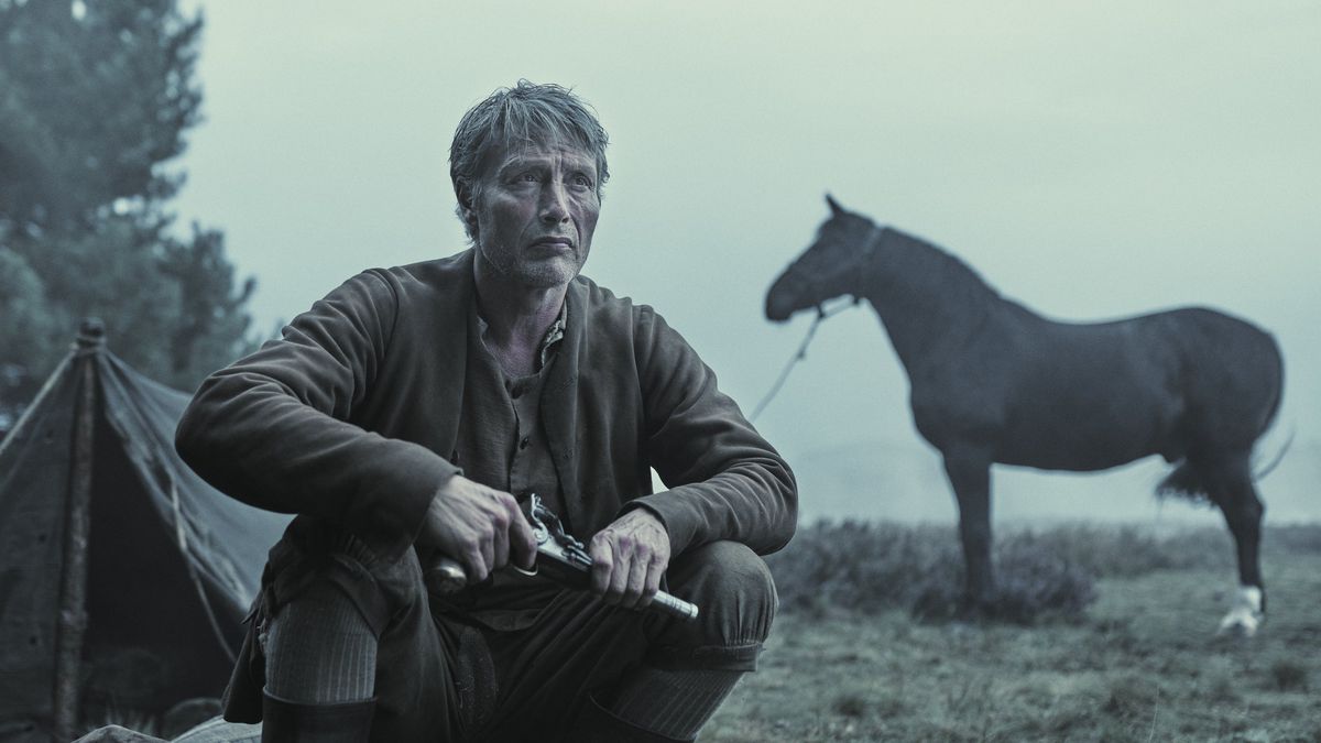 Mads Mikkelsen hält einen altmodischen Revolver in der Hand, während er in „Das gelobte Land“ neben einem Pferd und einem Zelt kniet