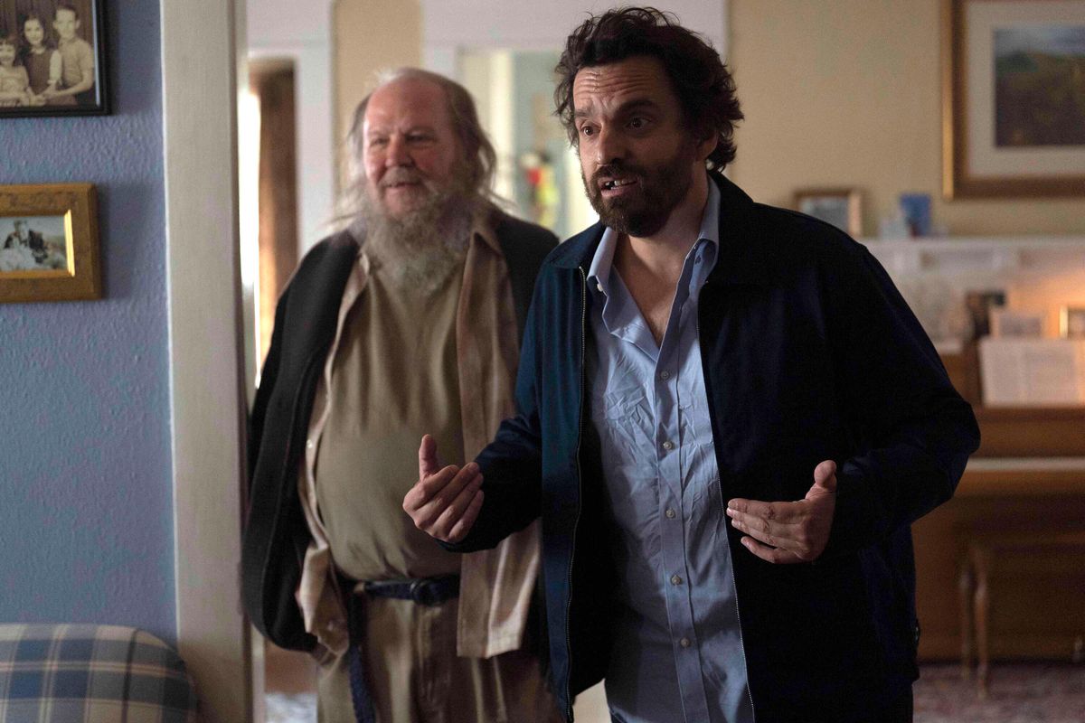 Tommy (Jake Johnson) and a friend (Biff Wiff) stand together in a living room, with Tommy earnestly telling a story to someone offscreen in Self Reliance