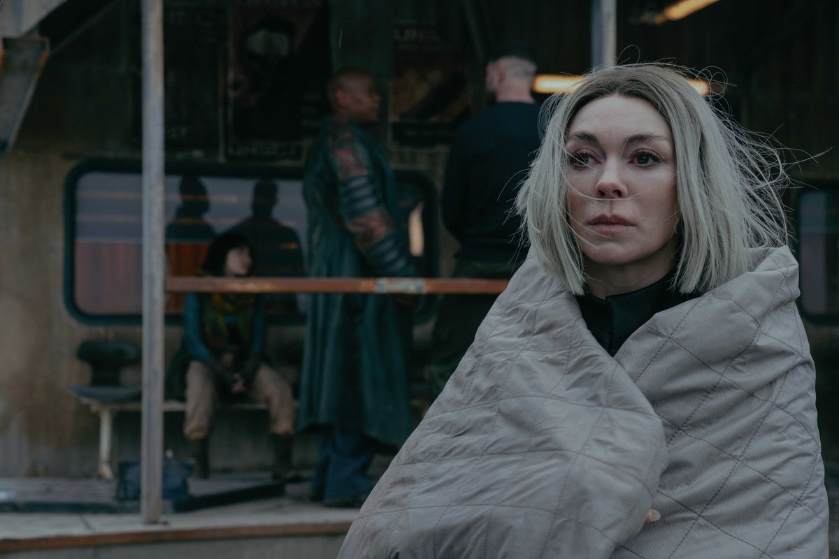 Fiona O'Shaughnessy como Laera en la temporada 2 de Halo está envuelta en una manta con dos personas hablando detrás de ella en un porche.
