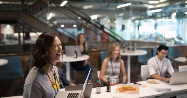 Empleado riendo sosteniendo una computadora portátil con compañeros de trabajo en segundo plano alrededor de una mesa de oficina