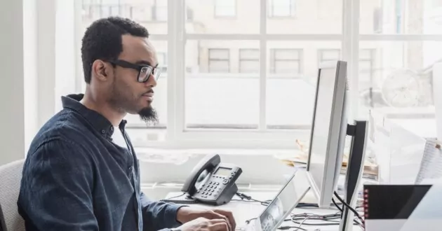 شخص يرتدي نظارة ولحية صغيرة على المكتب ويكتب على الكمبيوتر المحمول وينظر إلى الشاشة