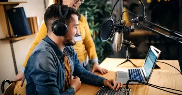 Dos compañeros de trabajo sonriendo frente a la computadora, uno sentado en una silla y usando auriculares, el otro de pie