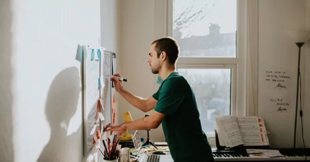 A man leans over a messy desk and writes on a wall-mounted whiteboard in a home-office environment