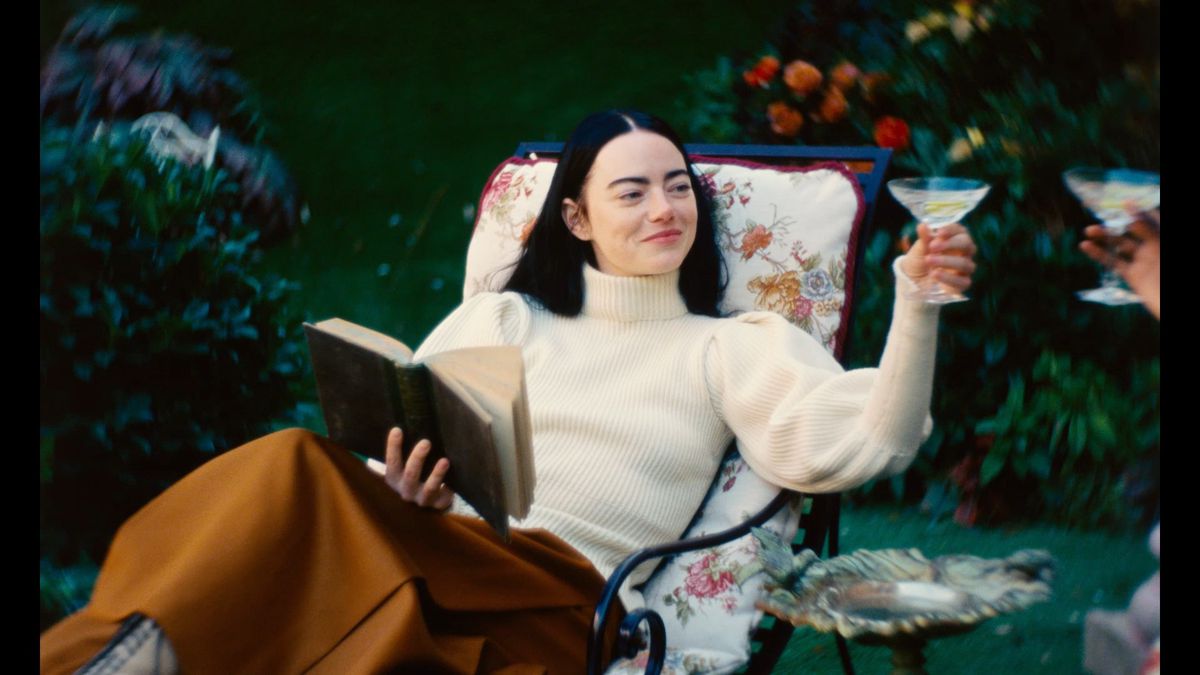 पूअर थिंग्स में बेला बैक्सटर के रूप में एम्मा स्टोन एक बगीचे की कुर्सी पर आराम कर रही हैं, एक किताब पकड़े हुए हैं, मुस्कुरा रही हैं और कॉकटेल का गिलास उठा रही हैं।