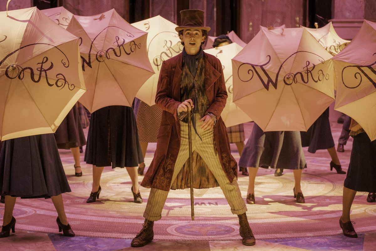 Wonka steht mit seinem Stock vor sich, während Tänzer im Film Wonka Regenschirme mit der Aufschrift „Wonka“ hochhalten.