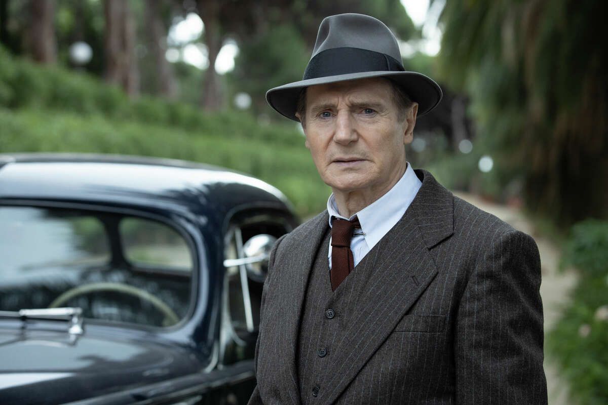 Een man (Liam Neeson) die in een bosrijk gebied staat voor een donkere sedanjurk in een bruin krijtstreeppak, donkerrode stropdas en een grijze gleufhoed.