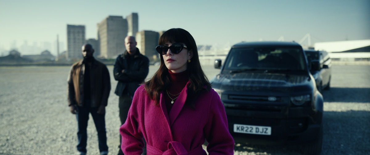 수지 글래스 역의 카야 스코델라리오(Kaya Scodelario)는 영화 <젠틀맨>에서 분홍색 코트를 입고 차 앞에 서 있습니다.