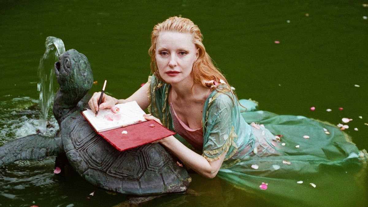 Shere Hite, gekleidet in ein hellgrünes Kleid, schwebt mit aufgeschlagenem Notizbuch auf einer Schildkrötenstatue im Wasser. Sie sieht mehr als nur ein wenig aus wie eine Meerjungfrau. Das Bild stammt aus der Dokumentation The Disappearance of Shere Hite