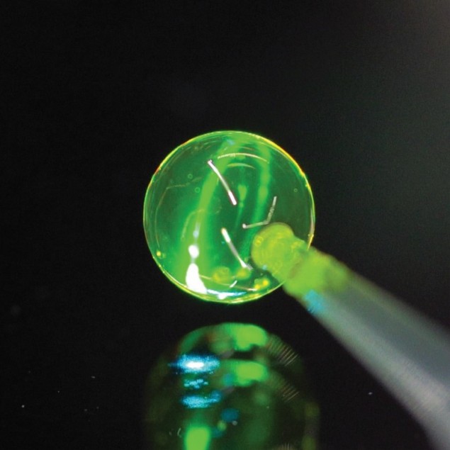 Foto de una pompa de jabón al final de un tubo capilar, bañada por una luz láser de color verde amarillento.