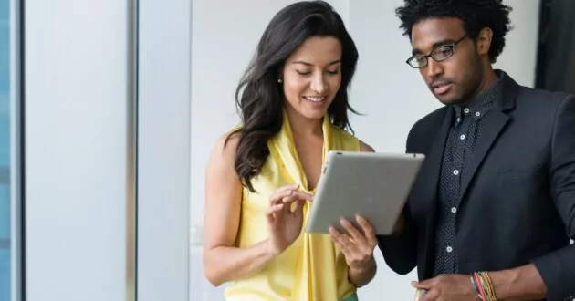 Zwei Geschäftsleute nutzen ein digitales Tablet im Büro, eine Person trägt ein gelbes Kleid, eine andere trägt einen Anzug