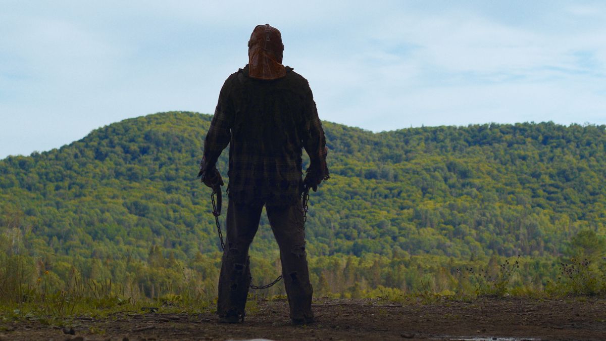 Uma figura com máscara e dois ganchos nas mãos fica de frente para uma floresta no filme In A Violent Nature