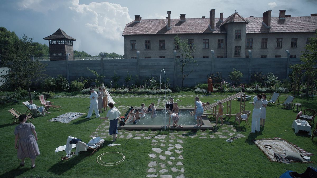 Varias personas se encuentran en un jardín amurallado con las torres de Auschwitz detrás de ellas en la Zona de Interés.