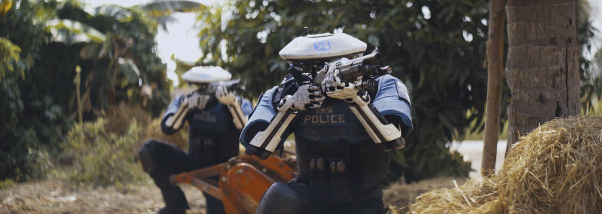 Tropikal bir bölgede çömelmiş iki robot polis memuru The Creator'a pompalı tüfeklerini doğrultuyor.