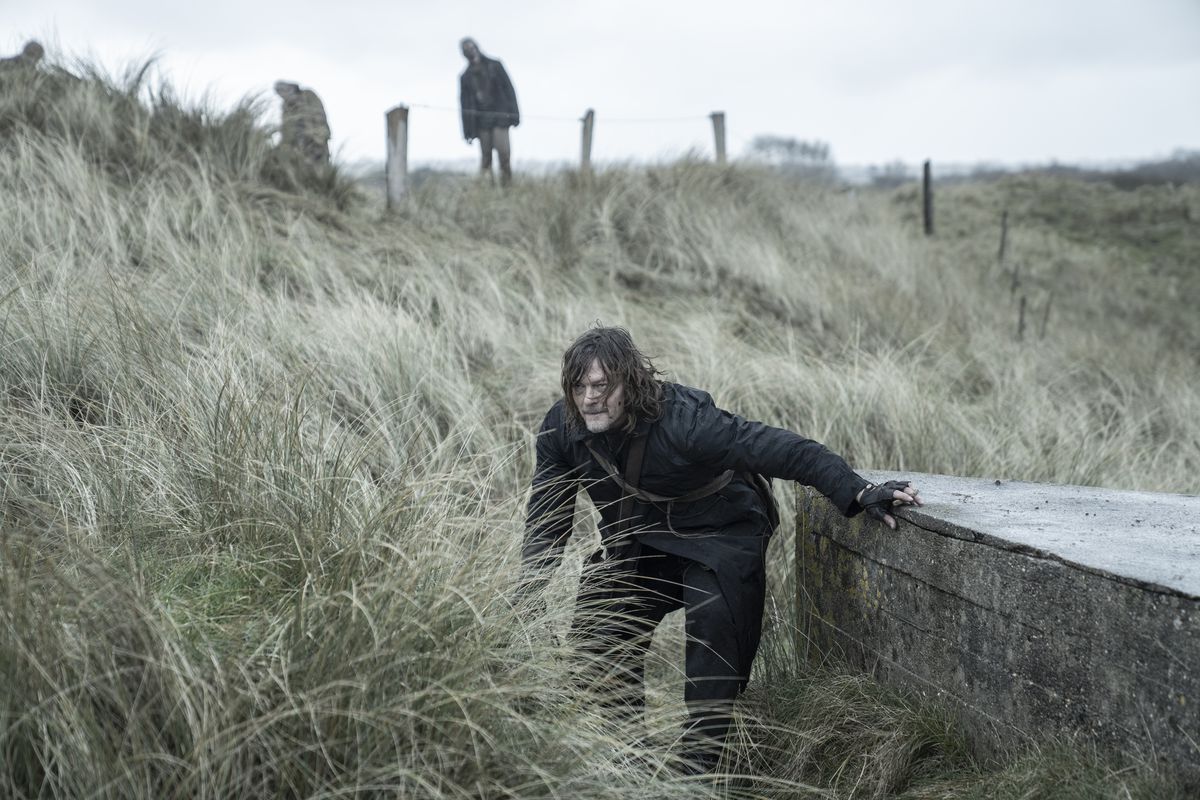 Daryl (Norman Reedus) agachado sobre la hierba alta con zombis al fondo detrás de él en una imagen fija de The Walking Dead: Daryl Dixon