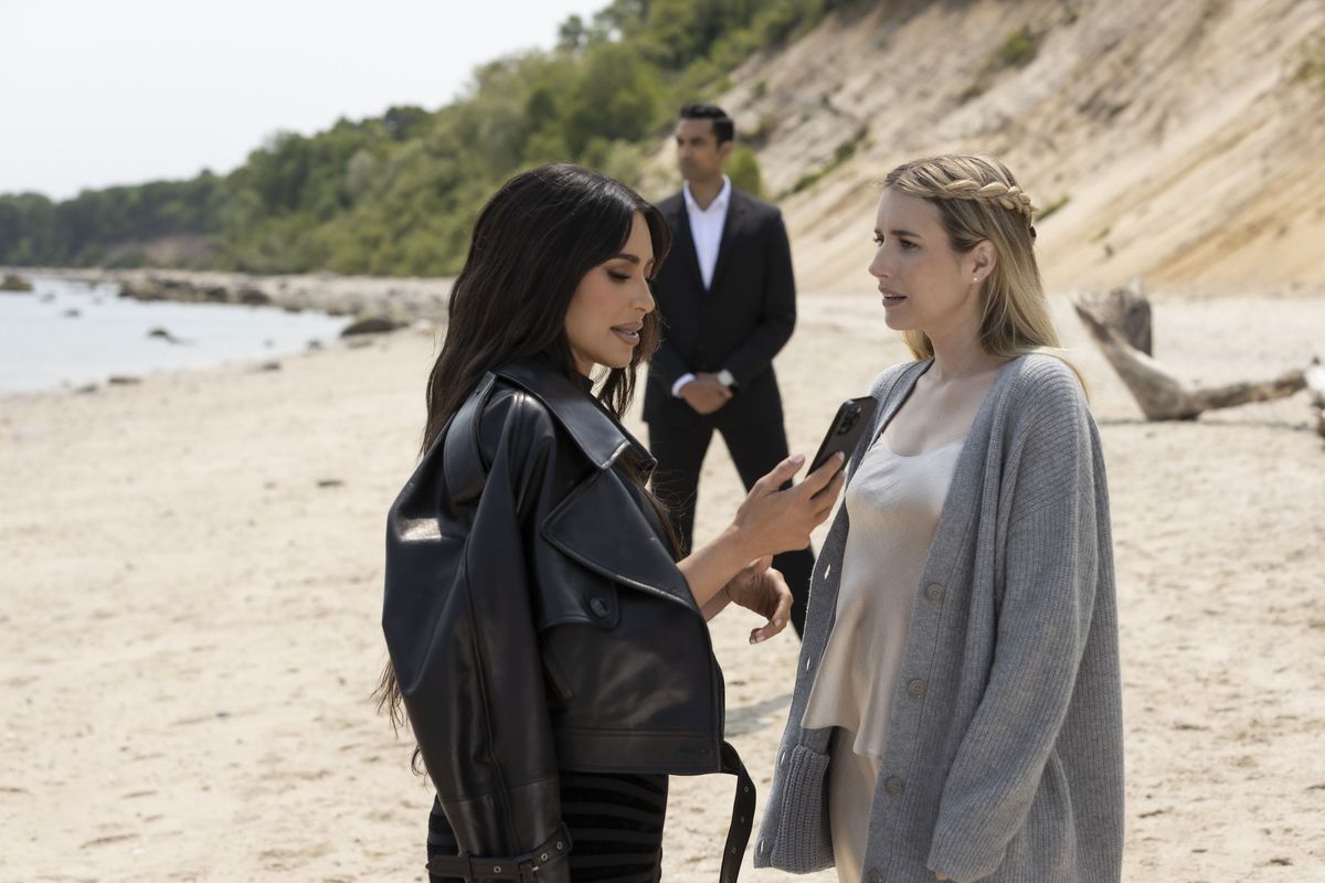 Siobhan (Kim Kardashian) kijkt naar haar telefoon terwijl Anna (Emma Roberts) met haar praat en vaag boos kijkt op een strand