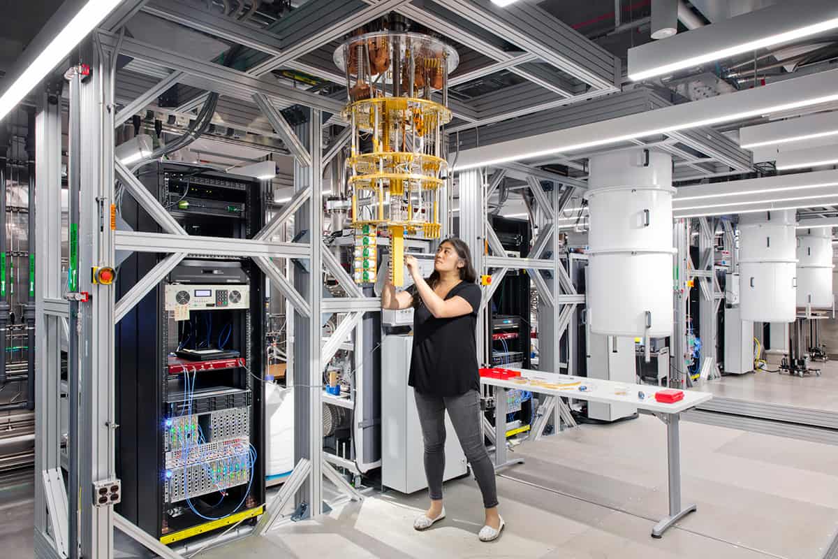 एक कंप्यूटिंग प्रयोगशाला जिसमें एक क्वांटम कंप्यूटर एक धातु के फ्रेम से लटका हुआ है और एक वैज्ञानिक इसके आधार पर कुछ समायोजित कर रहा है
