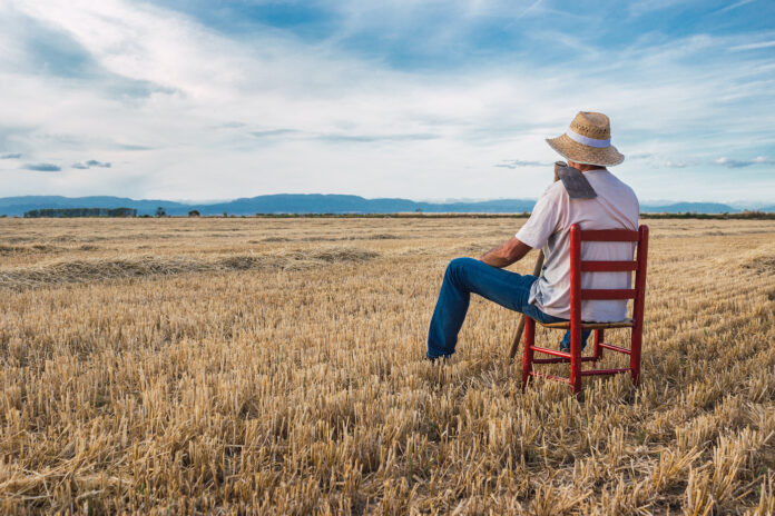 Farmer with straw hat, white t-shirt, and blue jeans sitting on a red chair surveying his land with a till on his shoulder