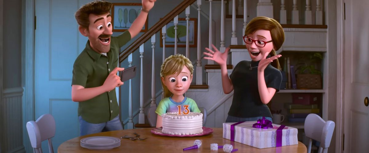Riley sits at a table looking at a birthday cake with “13” spelled out on it in candles as her mother and father cheer for her in Pixar Animation Studios’ Inside Out 2