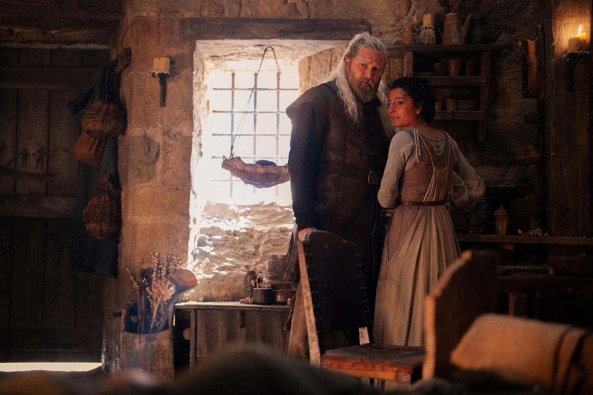 Kieran Bew as Hugh Hammer and Ellora Torchia as Hugh’s wife in House of the Dragon season 2 standing in a dimly lit room looking toward a door