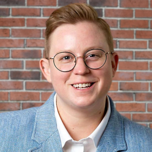 A headshot of Rae Lymer (they, them), who has short red hair and is wearing glasses and a blue blazer - and standing in front of a brick wall. 